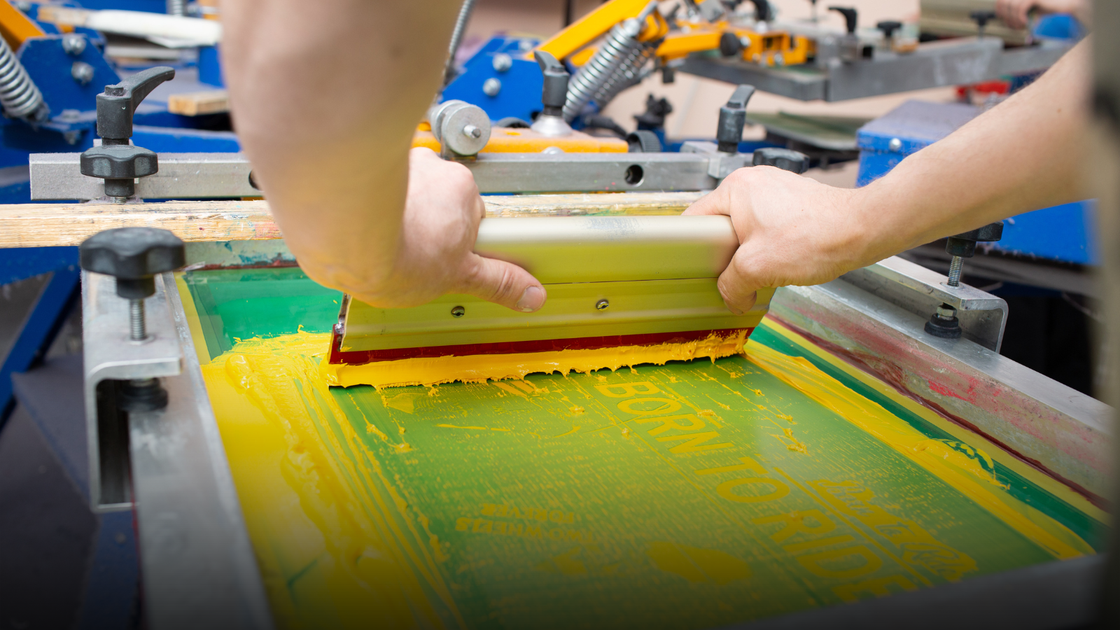 Person operating a screen printing press with a green screen and yellow ink.