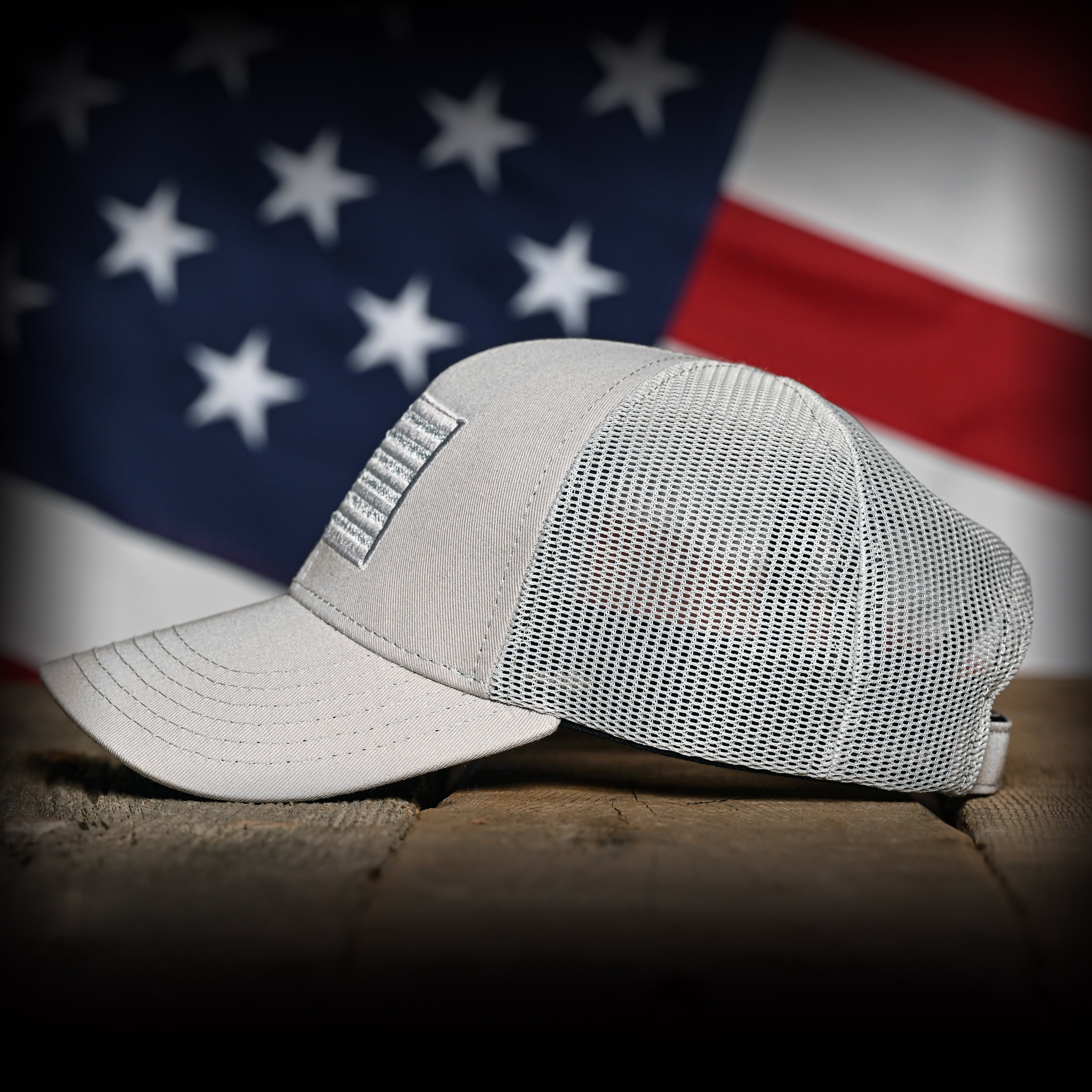 Left side view of a Tonal Light Gray American Flag Hat on a wood surface against a flag backdrop