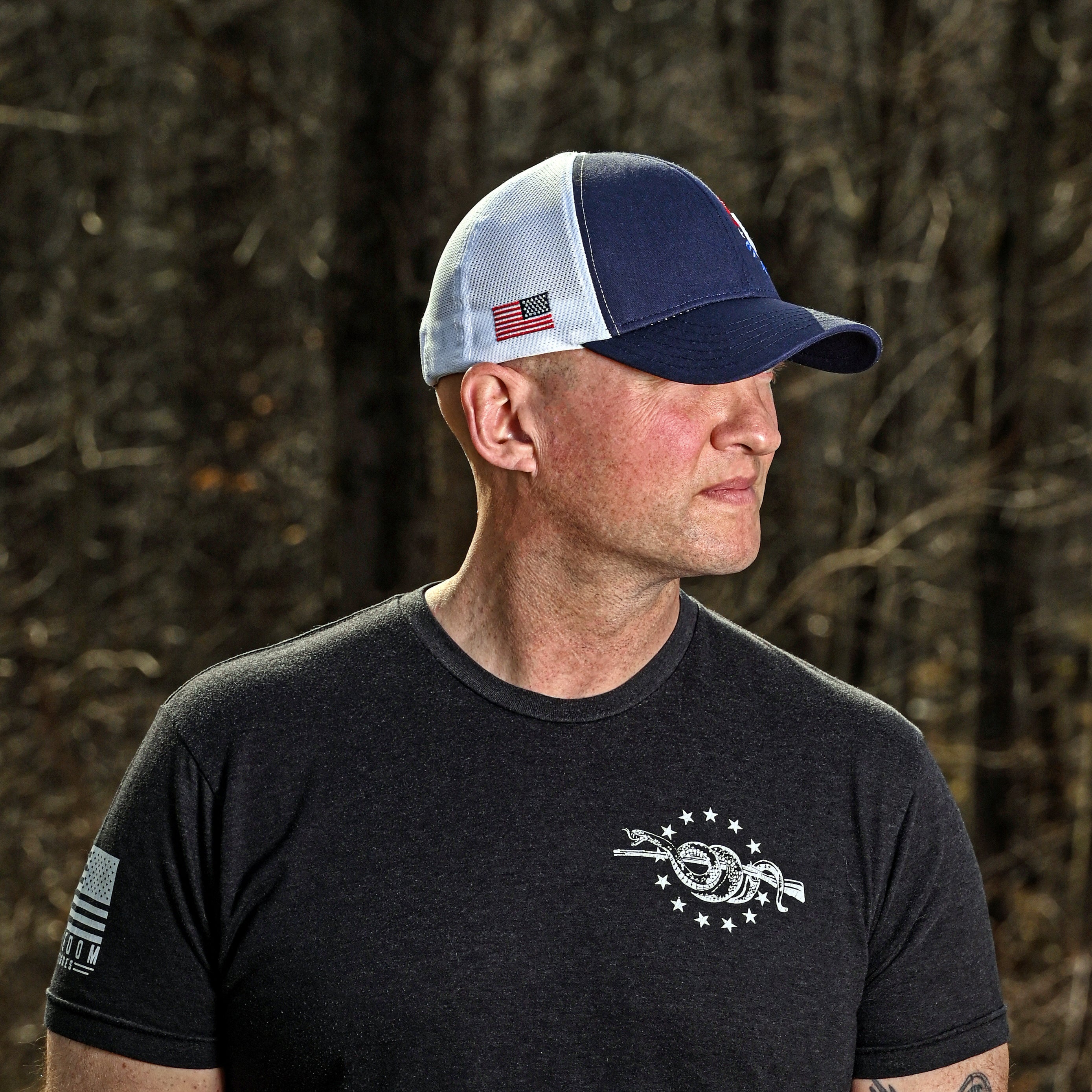Man wearing a black t-shirt with a logo and a cap, standing in a natural setting.