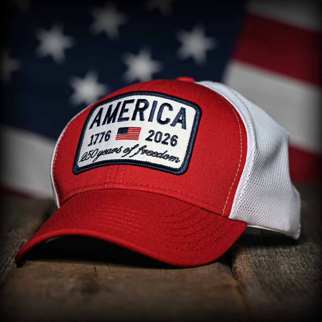 Red and white trucker hat with 'America 1776-2026' patch on a wooden surface with an American flag in the background.