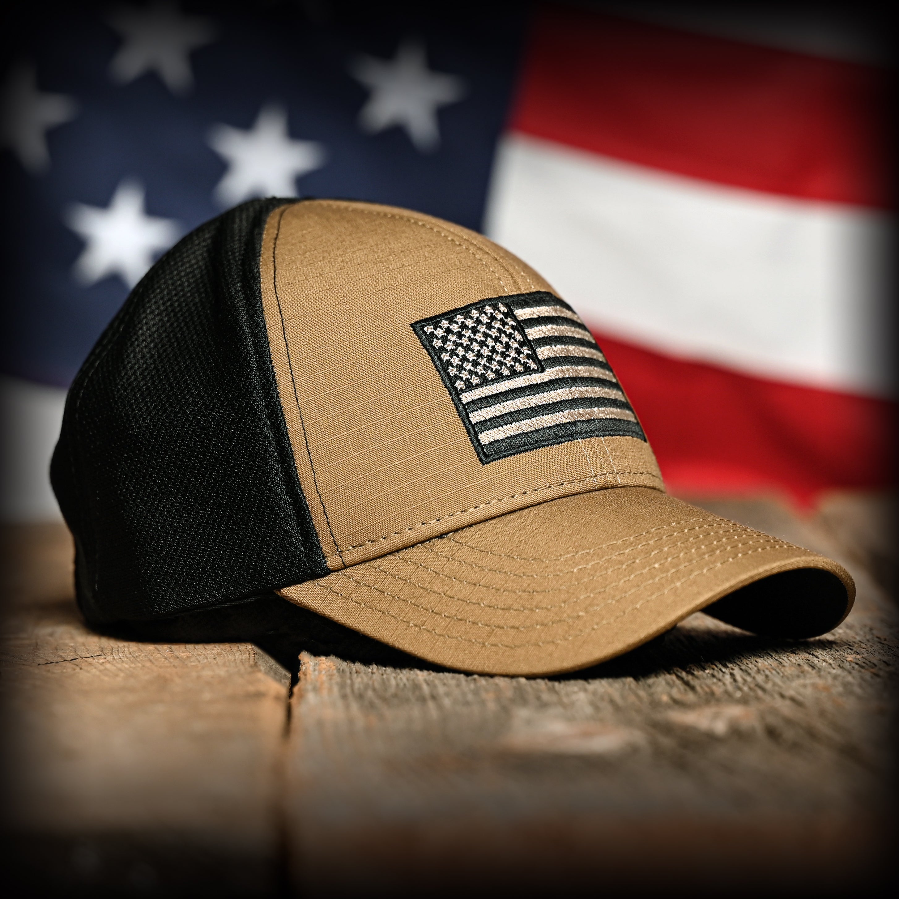 Front view of a coyote brown and black mesh cap embroidered with a flag on a wooden surface with an American flag in the background