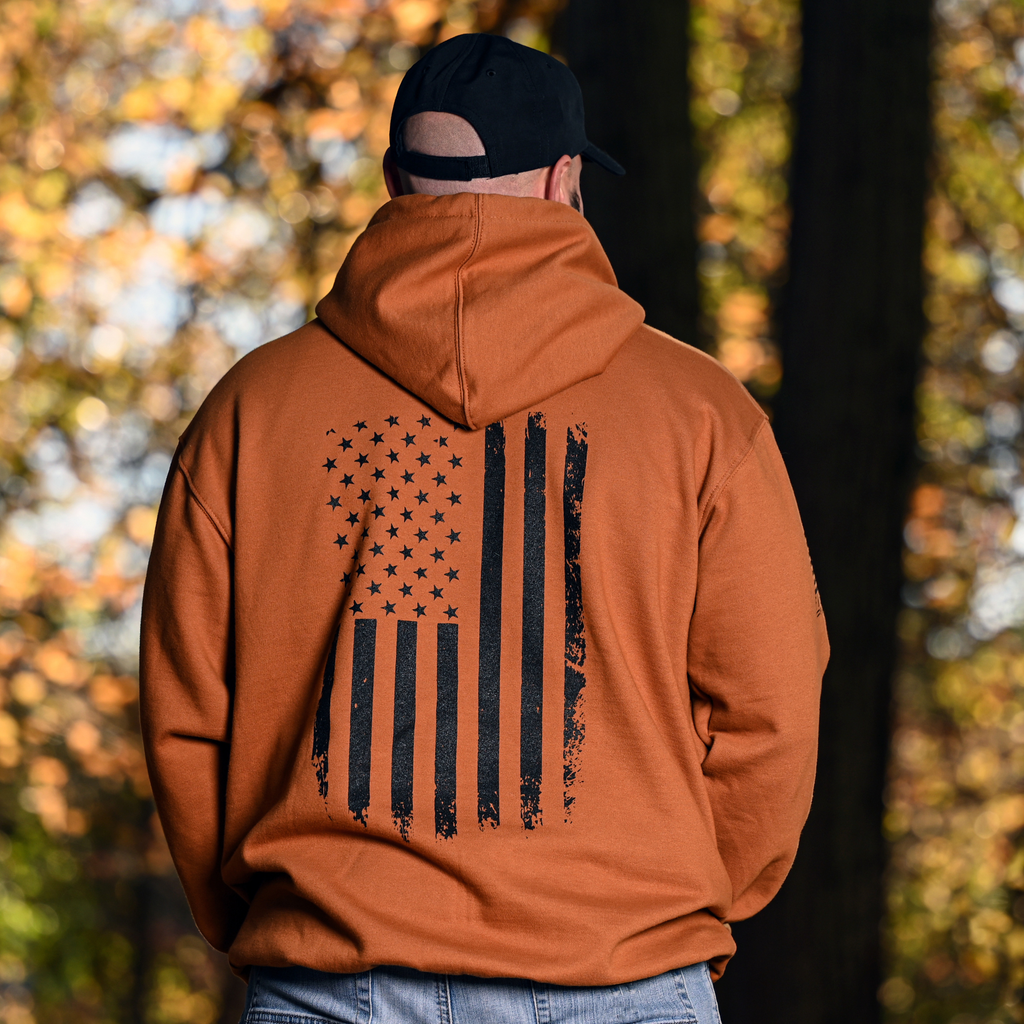 Man wearing a caramel brown hoodie with a black vertical American flag design in a forest setting