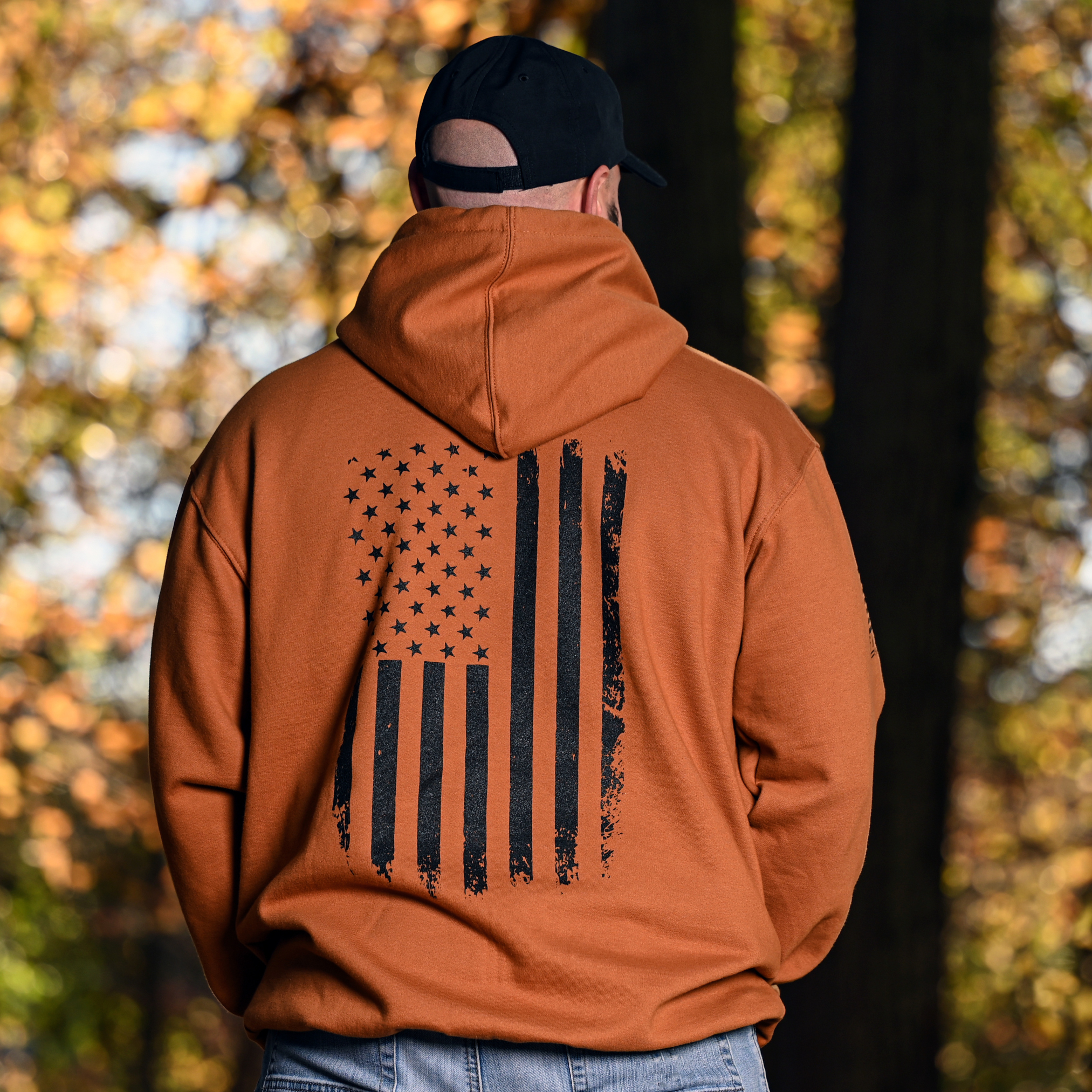Man wearing a caramel brown hoodie with a black vertical American flag design in a forest setting
