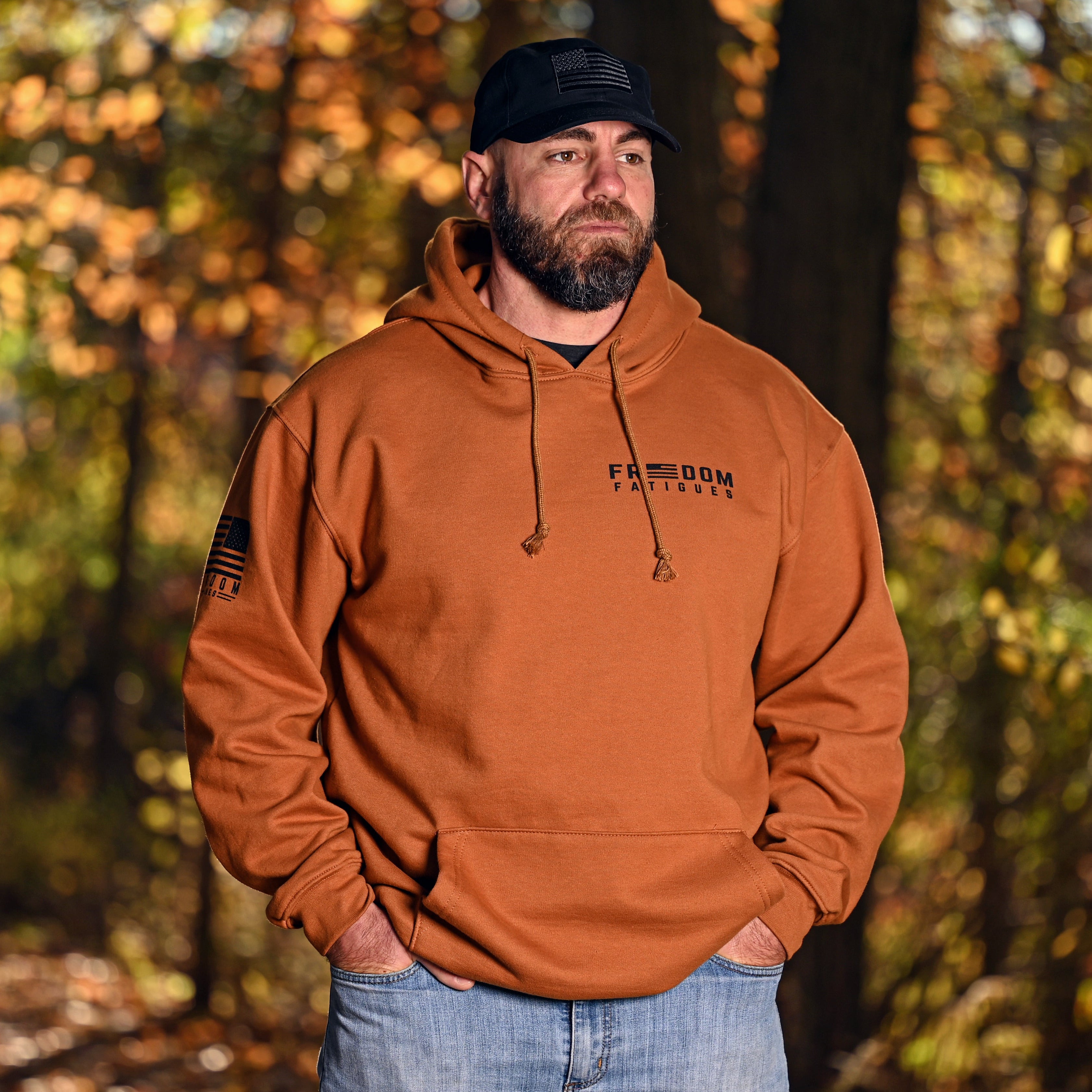 Man wearing a caramel brown hoodie with the Freedom Fatigues logo standing in a forest during autumn.