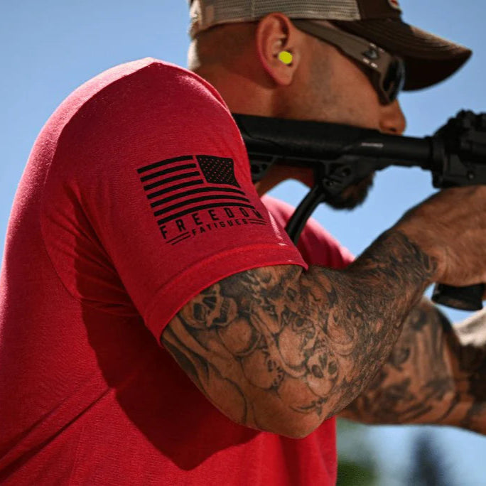 Person wearing a red t-shirt with a flag design, holding a rifle against a clear blue sky.
