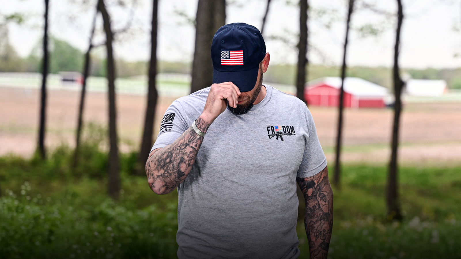 Man wearing the Freedom Stick t-shirt and a navy hat with an American flag, standing outdoors with trees and a barn in the background.