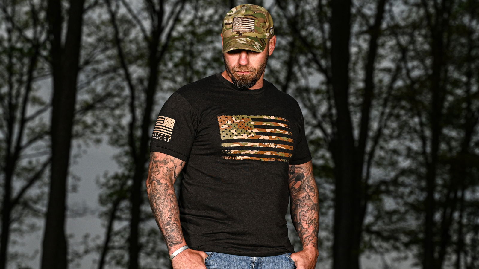 Man wearing a black t-shirt with an American flag design and a camouflage cap, standing against a forest background.