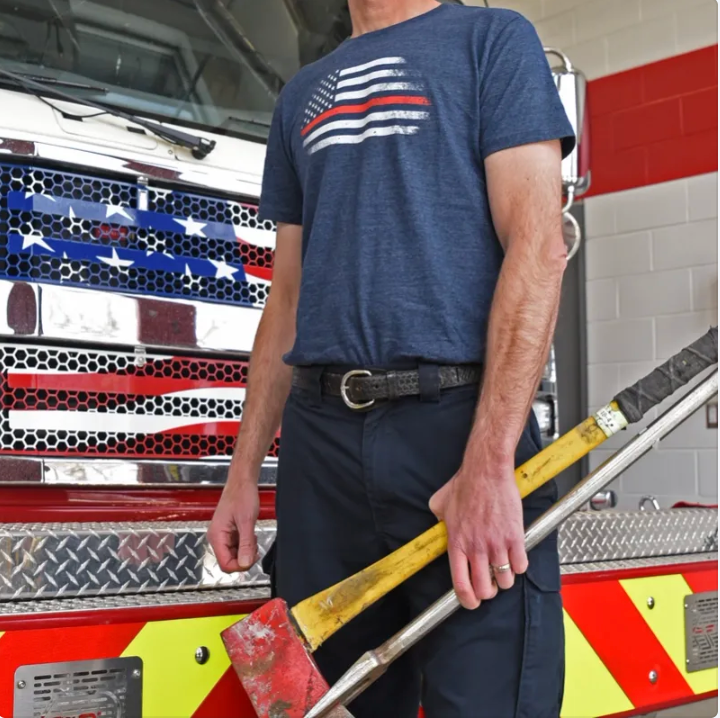 Fireman wearing a blue t-shirt with an thin red line American flag design, holding a fire axe in front of a fire truck.