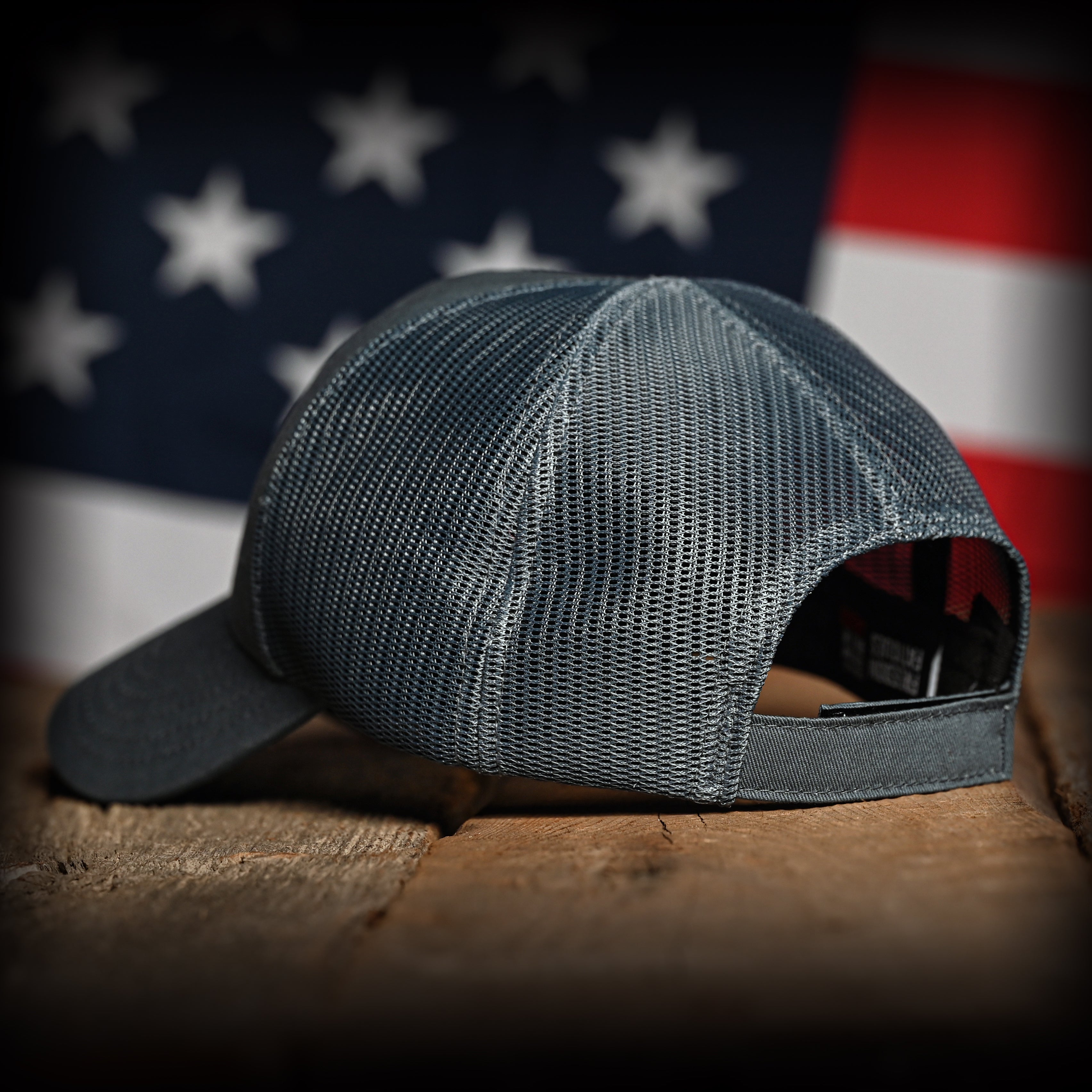 Tonal slate blue cap on a wooden surface with an American flag in the background, back left view