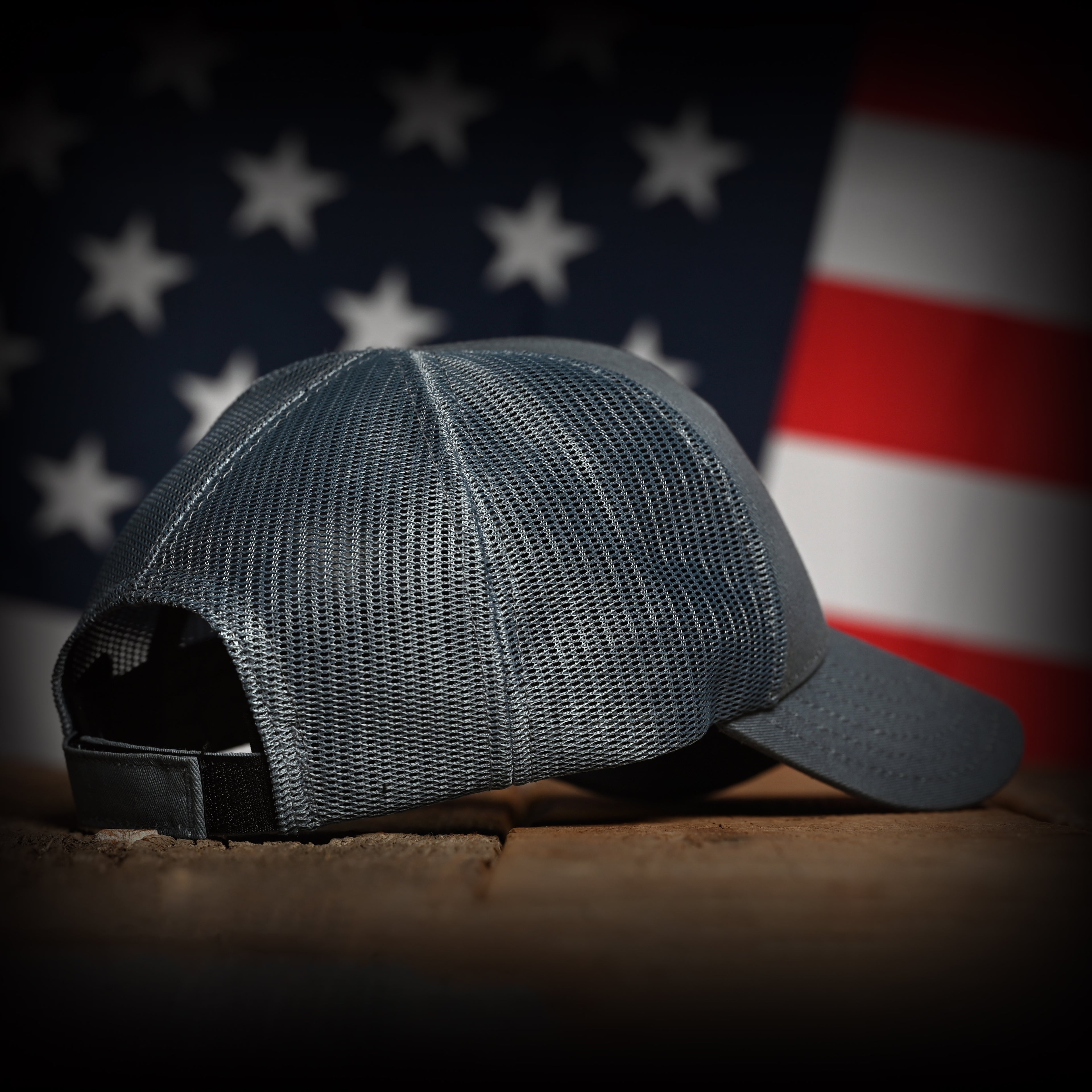 Tonal slate blue cap on a wooden surface with an American flag in the background, back right view
