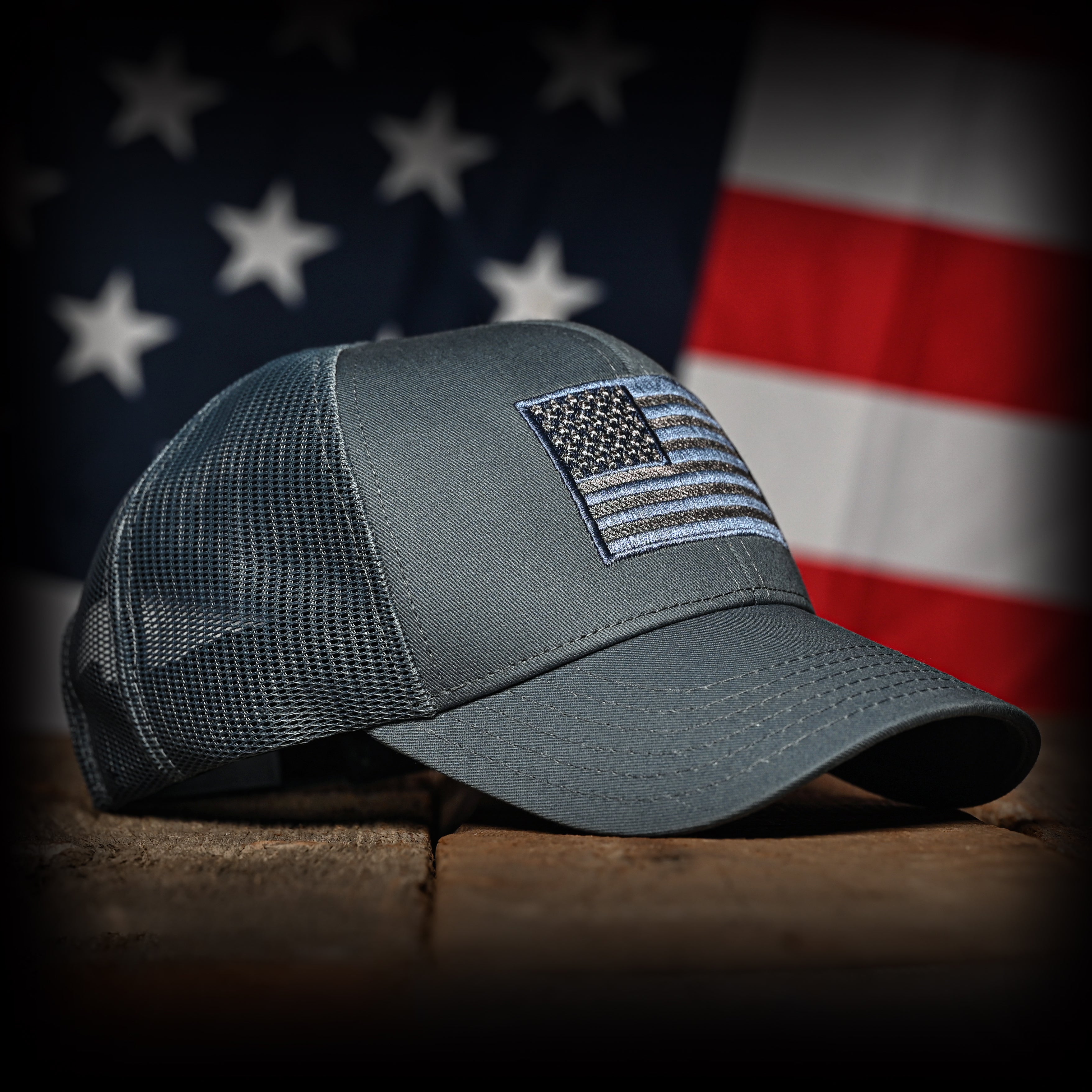 Tonal slate blue cap with American flag patch on a wooden surface with an American flag in the background, front right view