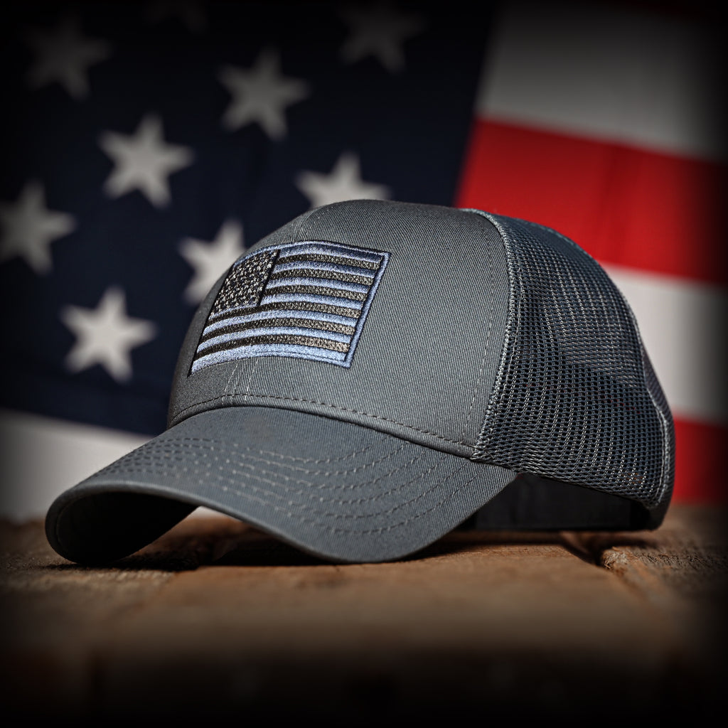 Tonal slate blue cap with American flag patch on a wooden surface with an American flag in the background