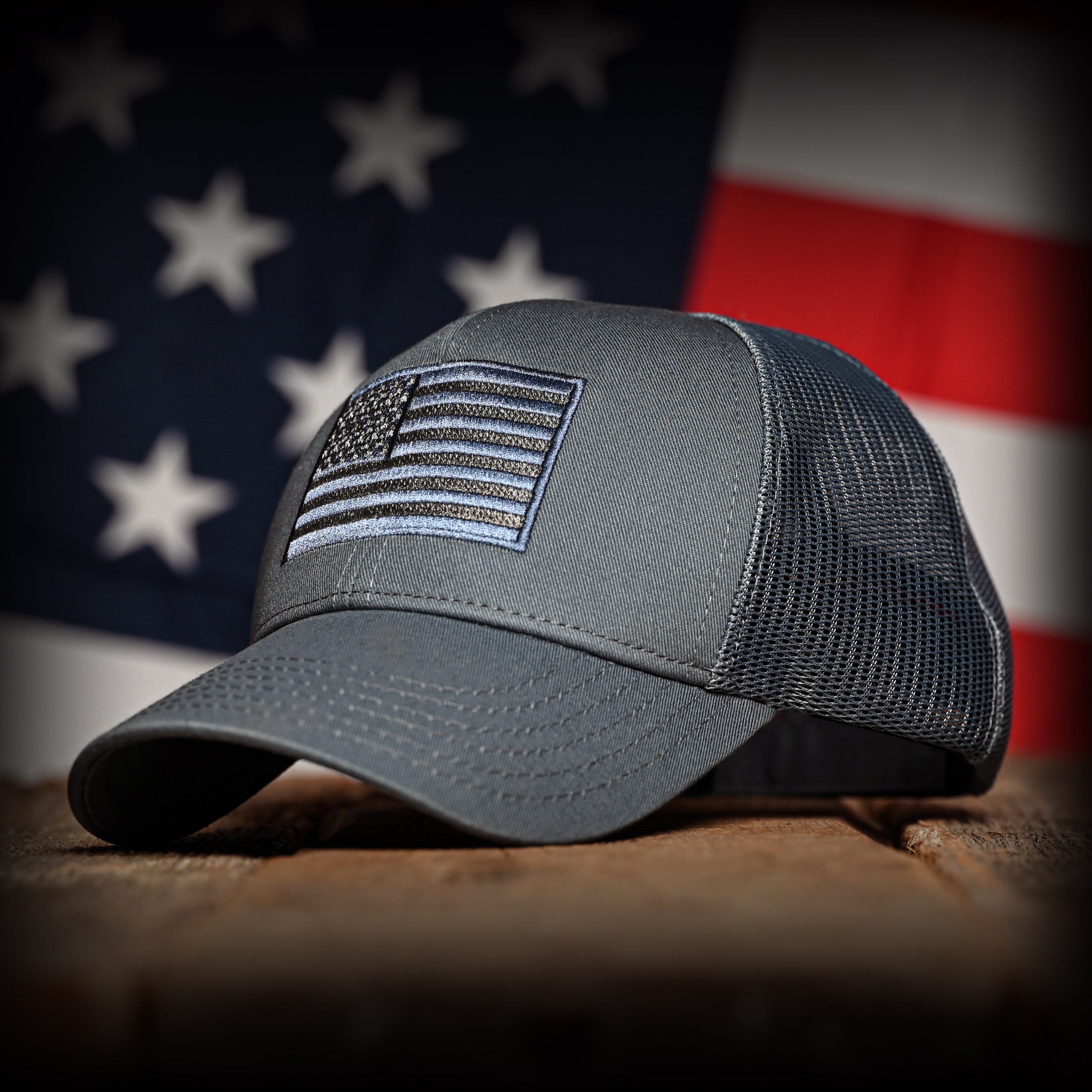 Tonal slate blue cap with American flag patch on a wooden surface with an American flag in the background