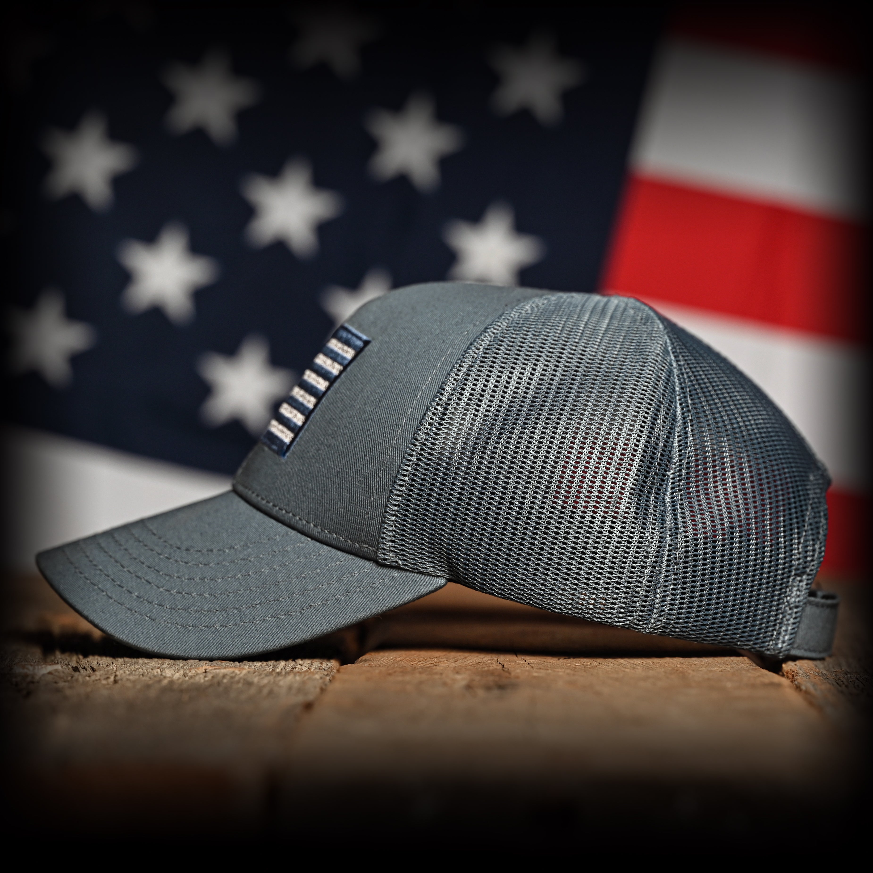 Tonal slate blue cap with an embroidered American flag on a wooden surface with an American flag in the background, left view