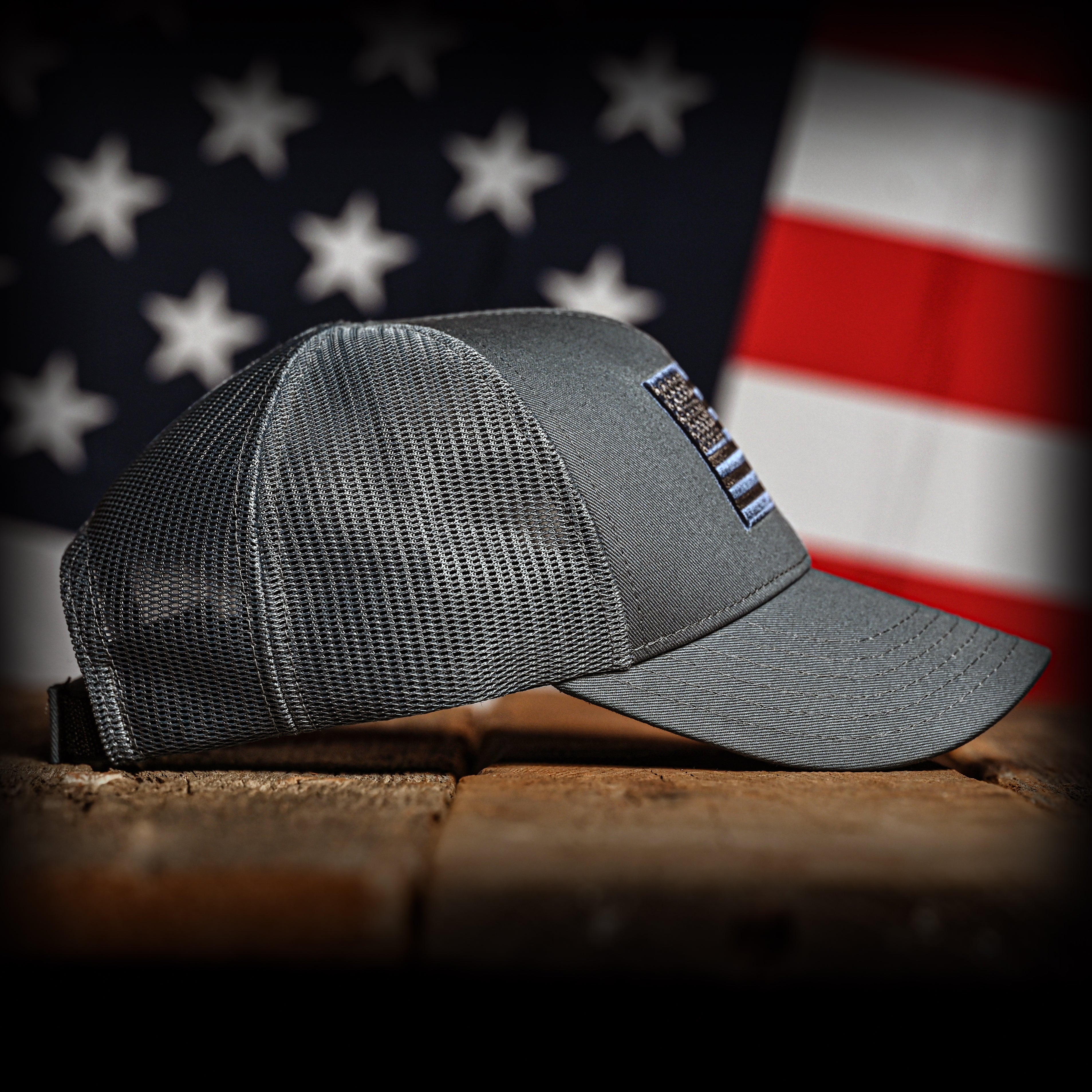 Tonal slate cap with an embroidered American flag on a wooden surface in front of an American flag, right view