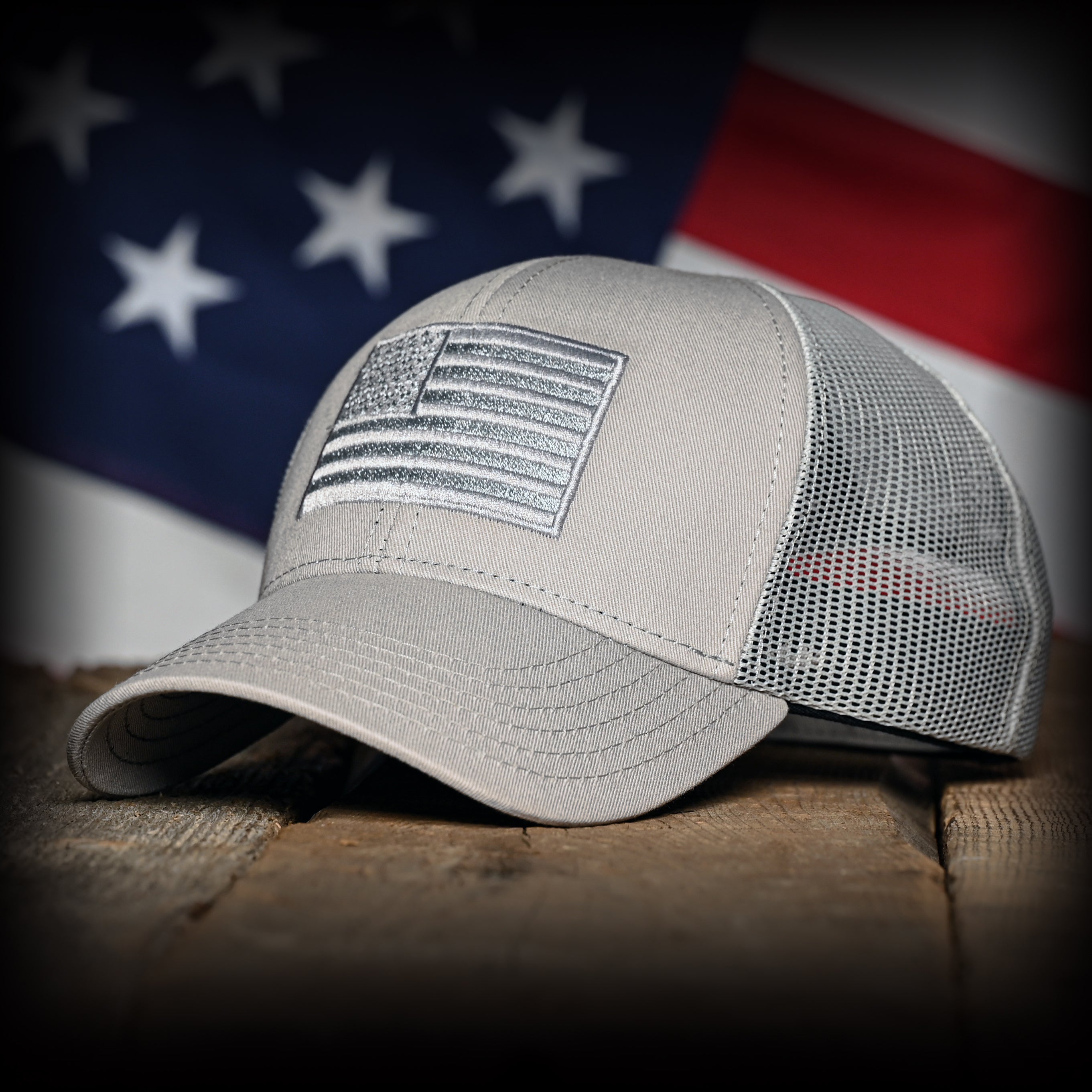 Tonal Light Gray American Flag Hat on a wood surface against a flag backdrop 