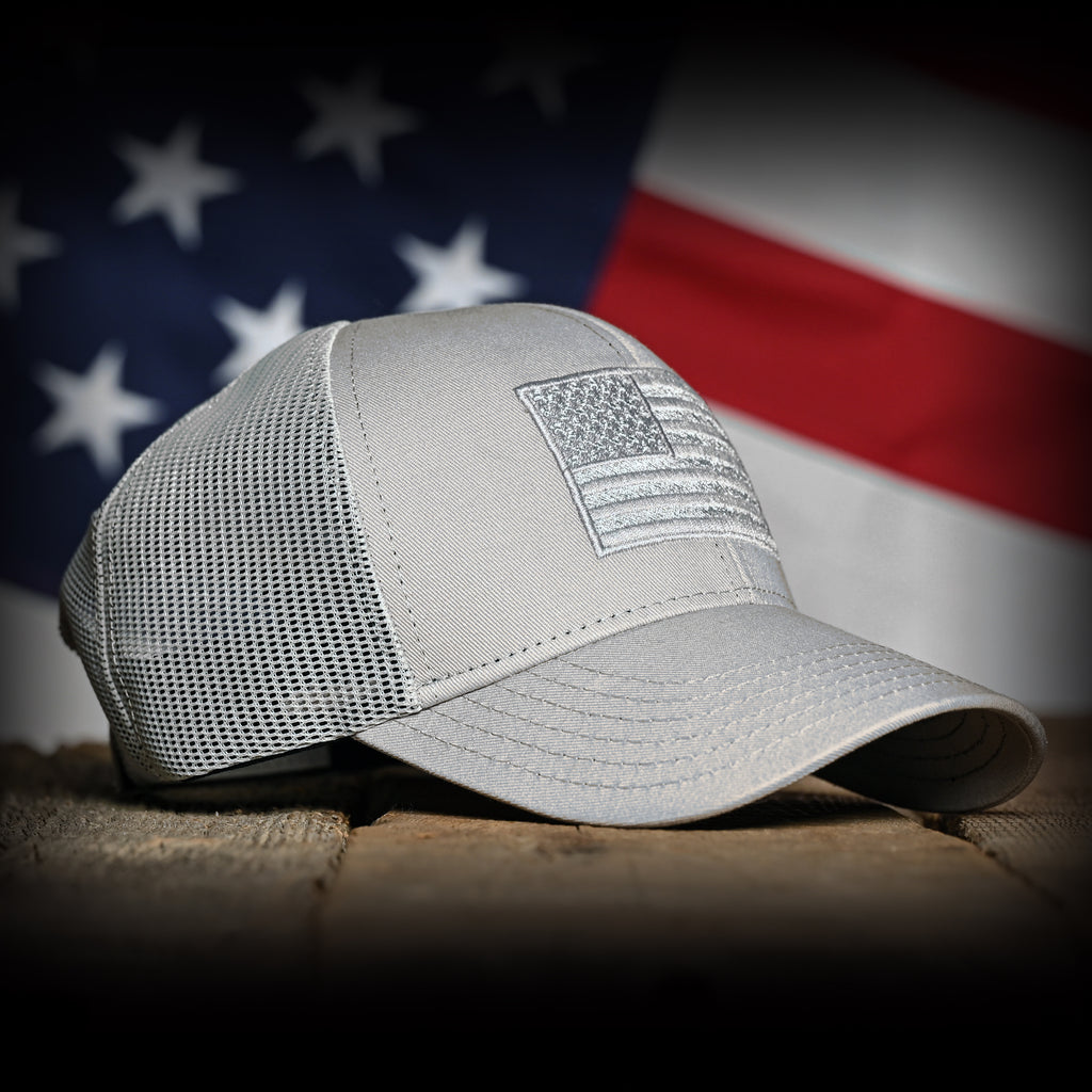 Front right view of a Tonal Light Gray American Flag Hat on a wood surface against a flag backdrop 