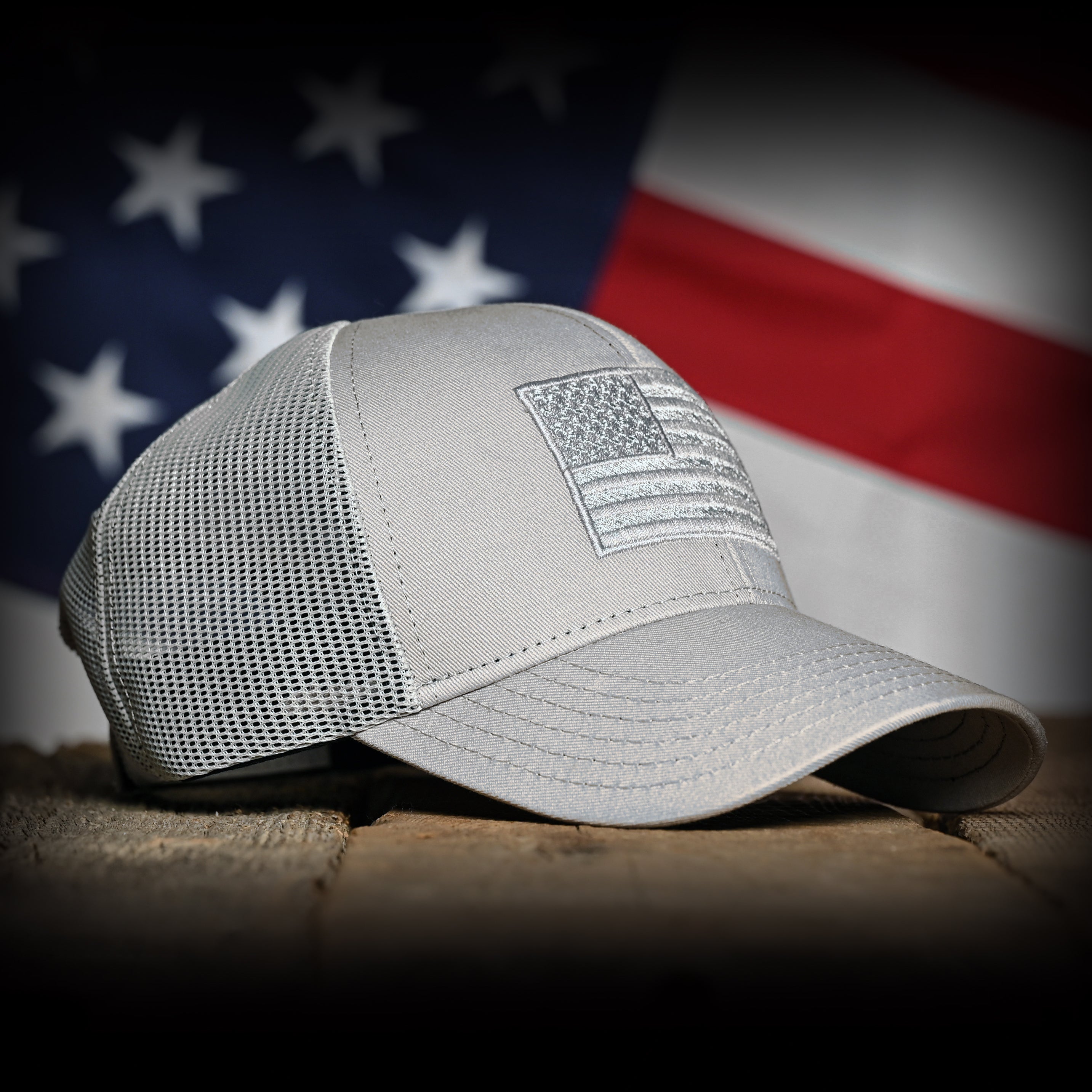Front right view of a Tonal Light Gray American Flag Hat on a wood surface against a flag backdrop 