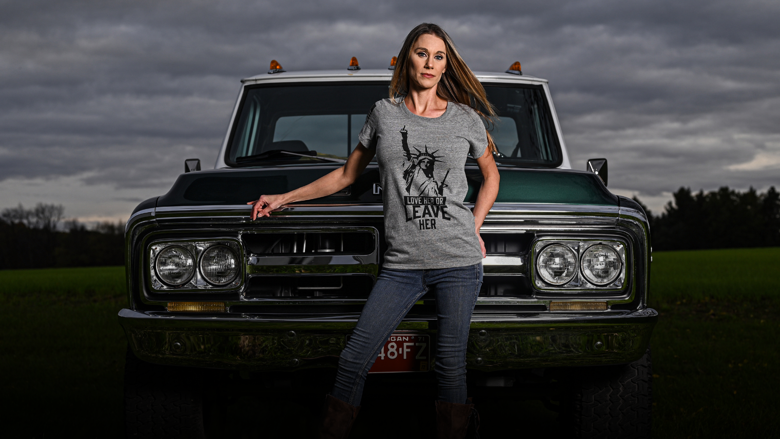 Woman wearing a patriotic tshirt standing in front of a vintage truck with a dark, moody sky.