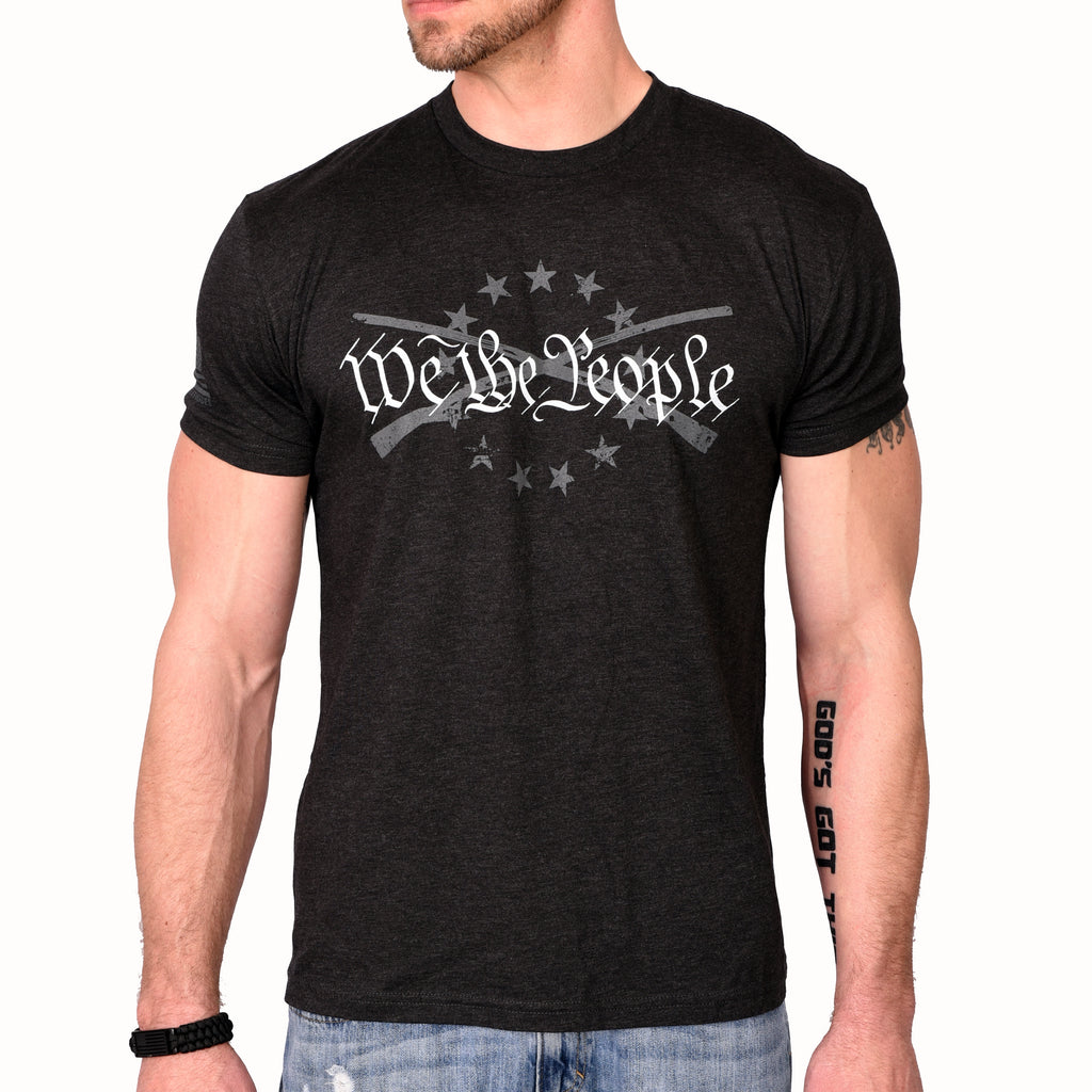 A male model wearing a black heather Men's T-shirt with the phrase 'We The People' printed in white, accompanied by graphics of crossed rifles and stars. (front view)