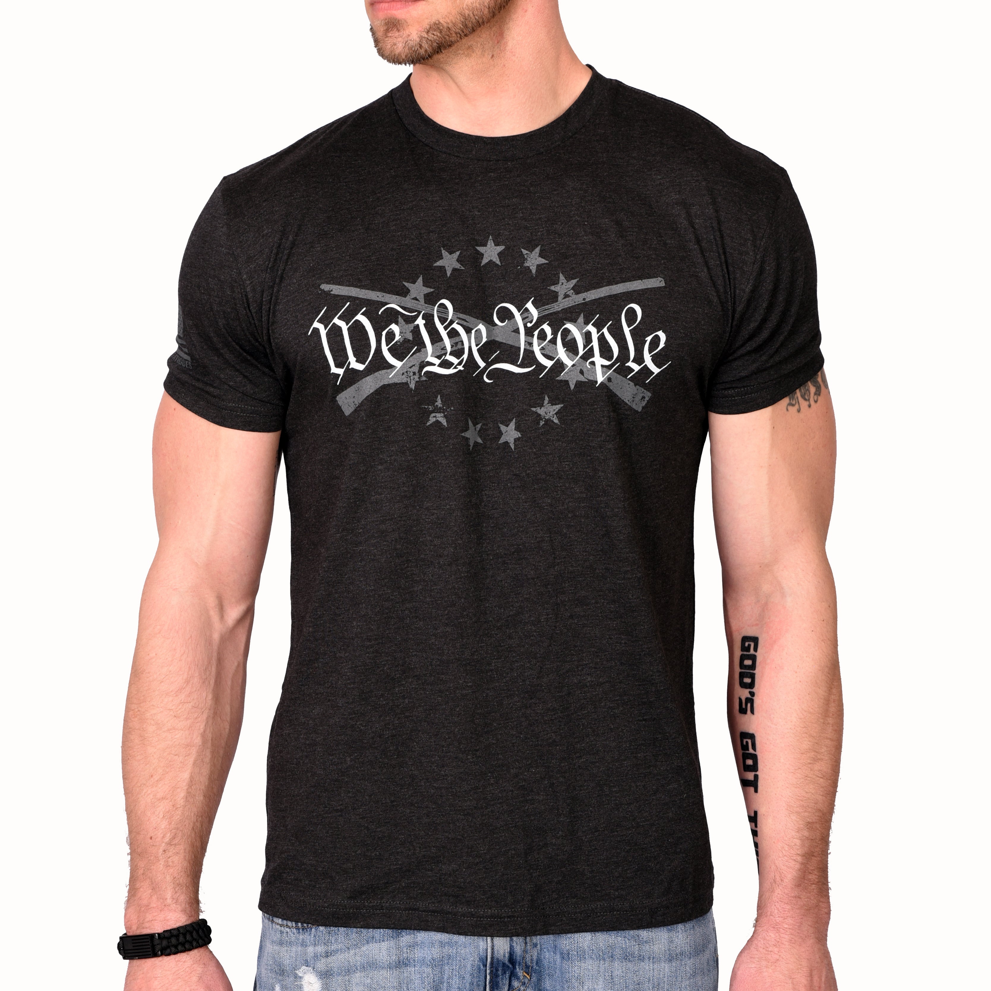 A male model wearing a black heather Men's T-shirt with the phrase 'We The People' printed in white, accompanied by graphics of crossed rifles and stars. (front view)