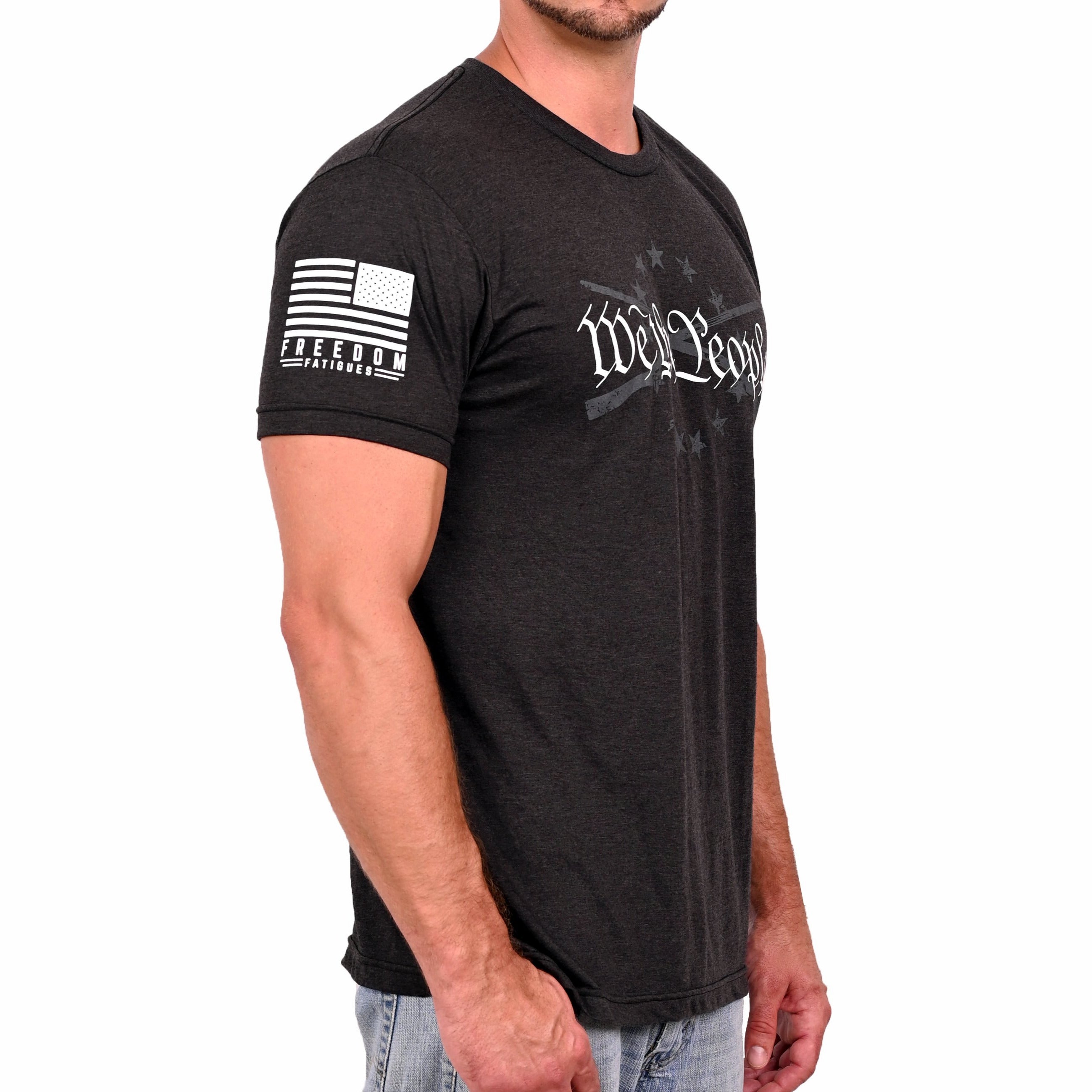 A male model wearing a black heather Men's T-shirt with the phrase 'We The People' printed in white, accompanied by graphics of crossed rifles and stars. (right view)